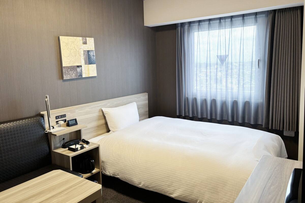 A hotel room with a large bed, a bedside table with a lamp, a small desk with a phone and a coffee maker, a headboard, and a window with curtains.