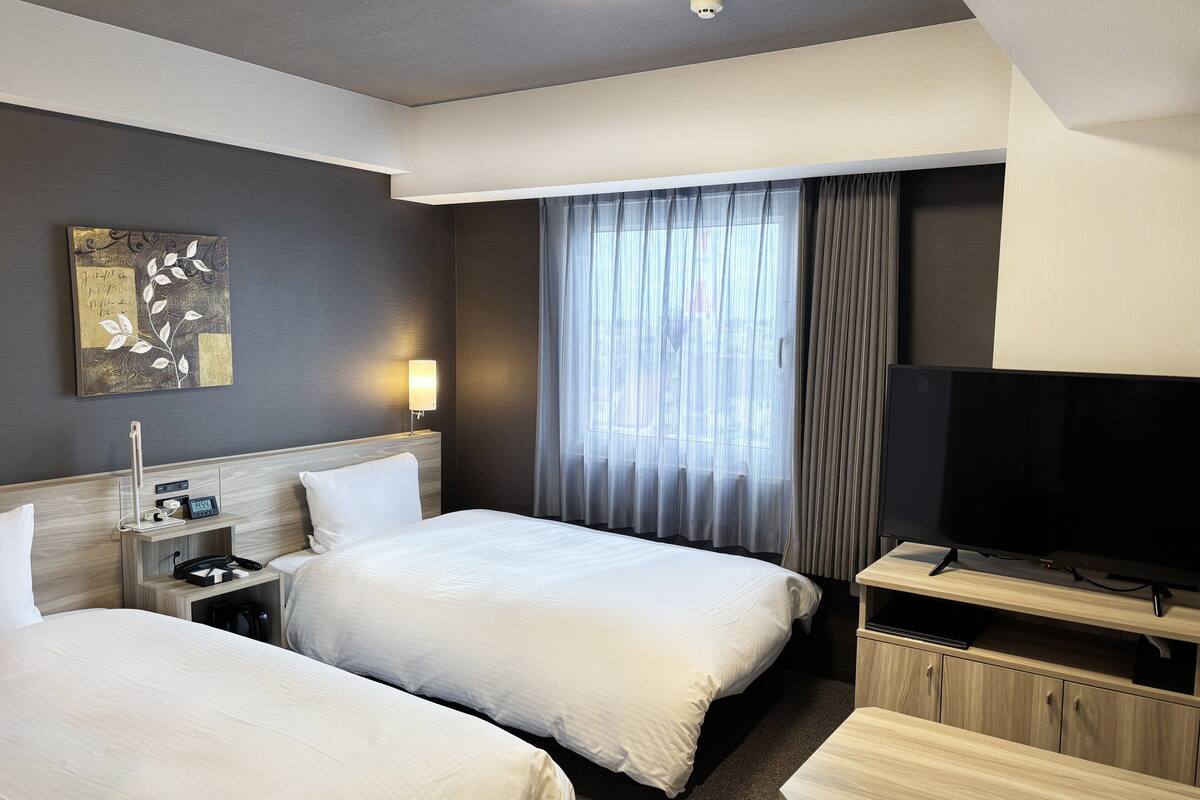 A hotel room with two beds, a bedside table with a lamp, a TV, and a painting on the wall.