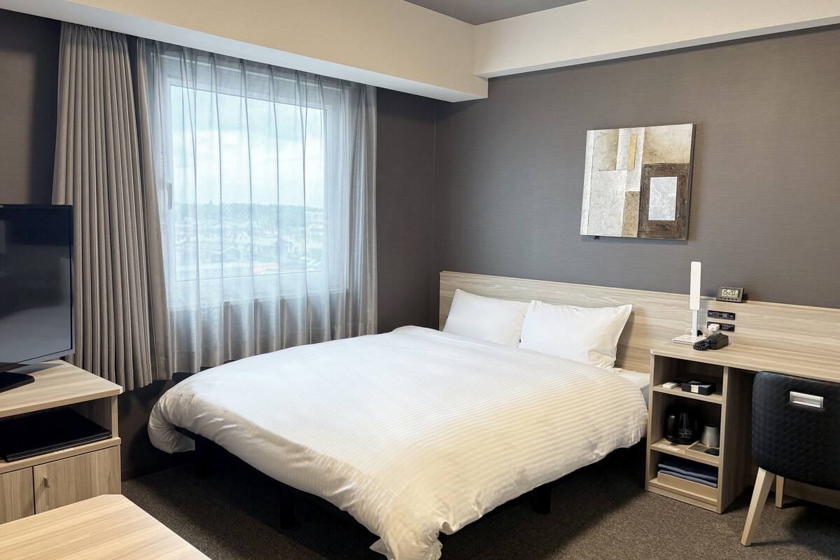 A hotel room with a bed, a desk, a TV, a nightstand, and a window with curtains.