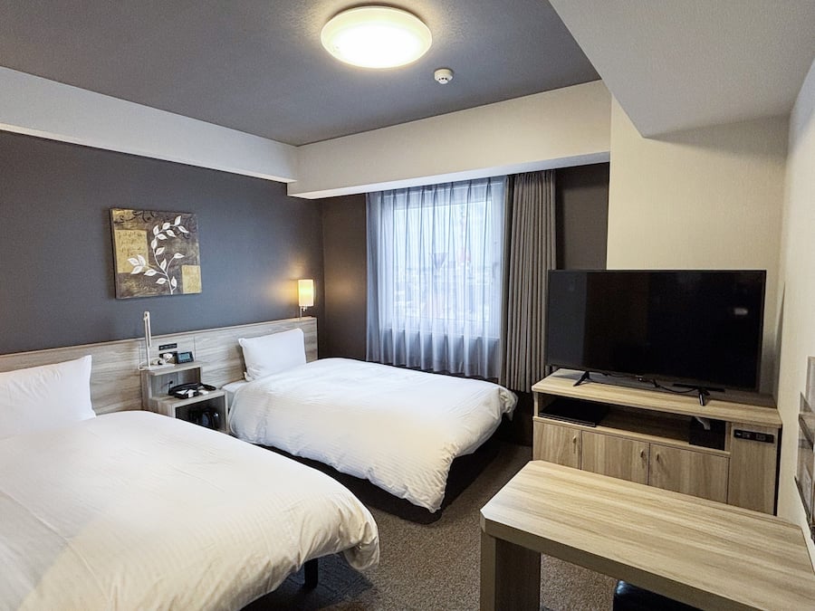 A hotel room with two beds, a TV, a desk, and a window with curtains.