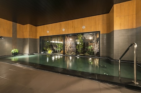 A modern indoor swimming pool with a wooden wall and glass doors, surrounded by plants and a metal railing.