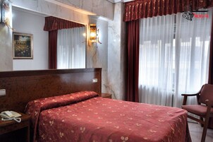 A hotel room with a bed, a chair, a bedside table with a phone, a mirror, and curtains.