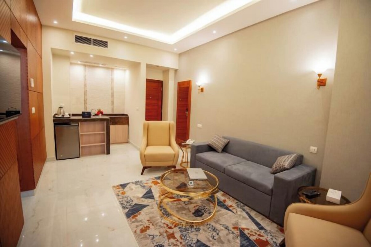 Una sala moderna con un sofá, sillones, una mesa de centro de vidrio y una cocina con refrigerador y alacenas.