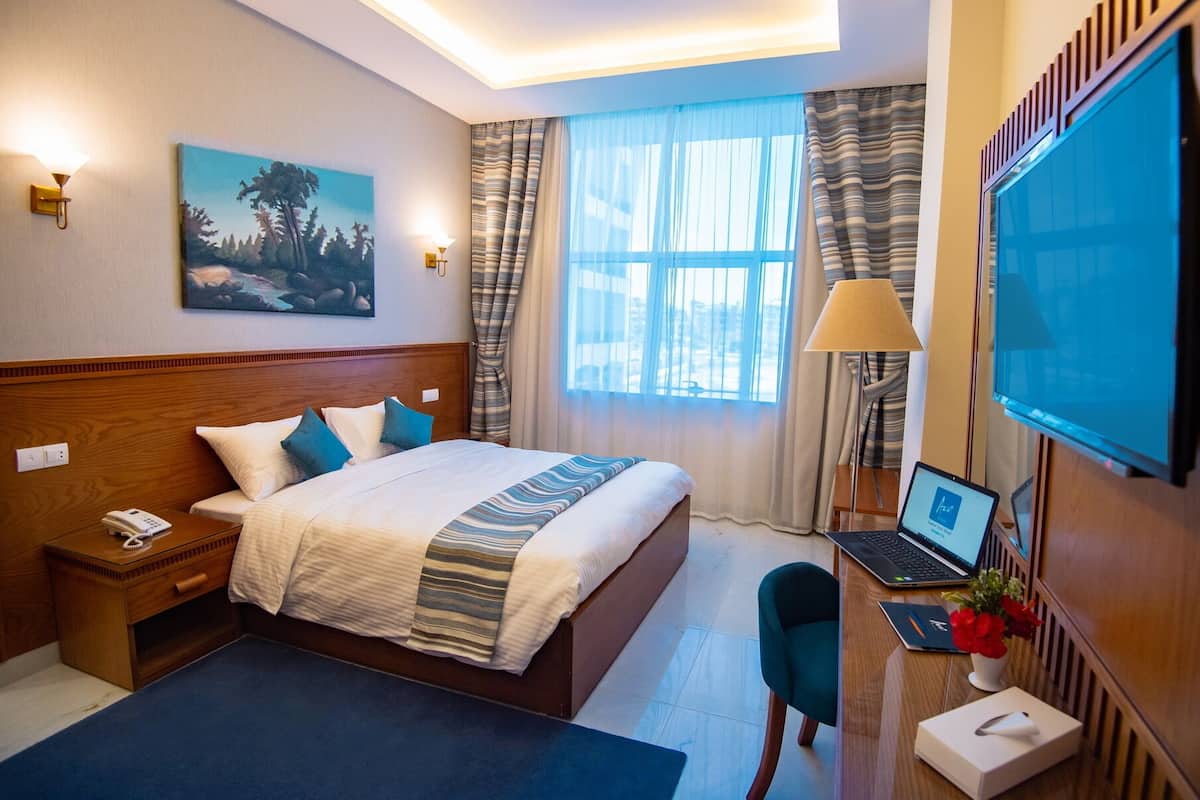 Habitación de hotel con cama, escritorio con una computadora portátil, televisor, un cuadro en la pared y ventana con cortinas.