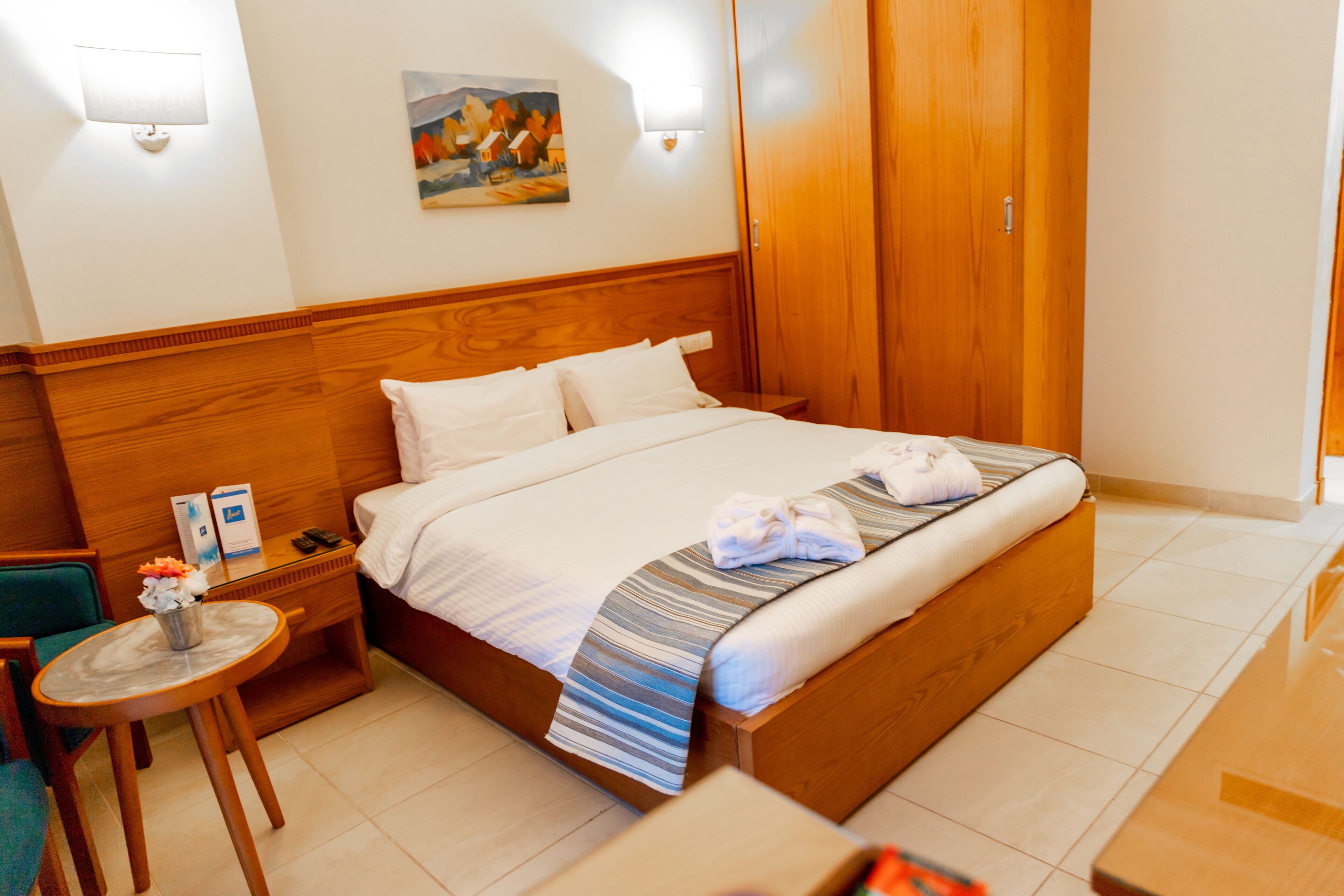 A hotel room with a large bed, a wooden headboard, a small round table with a vase of flowers, a nightstand with a lamp, and a painting on the wall.