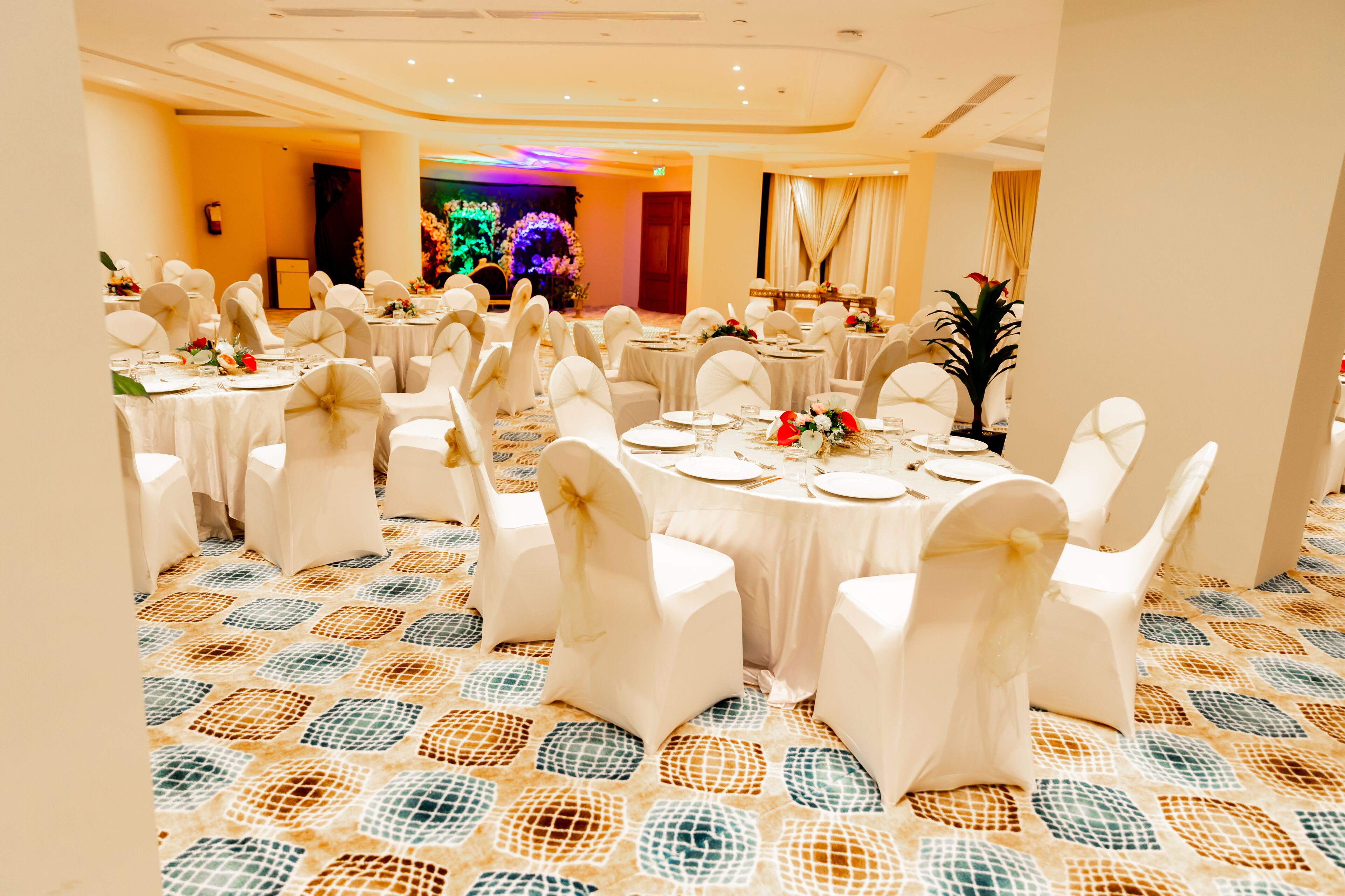Un salón de banquetes con mesas redondas, sillas blancas y un suelo estampado.