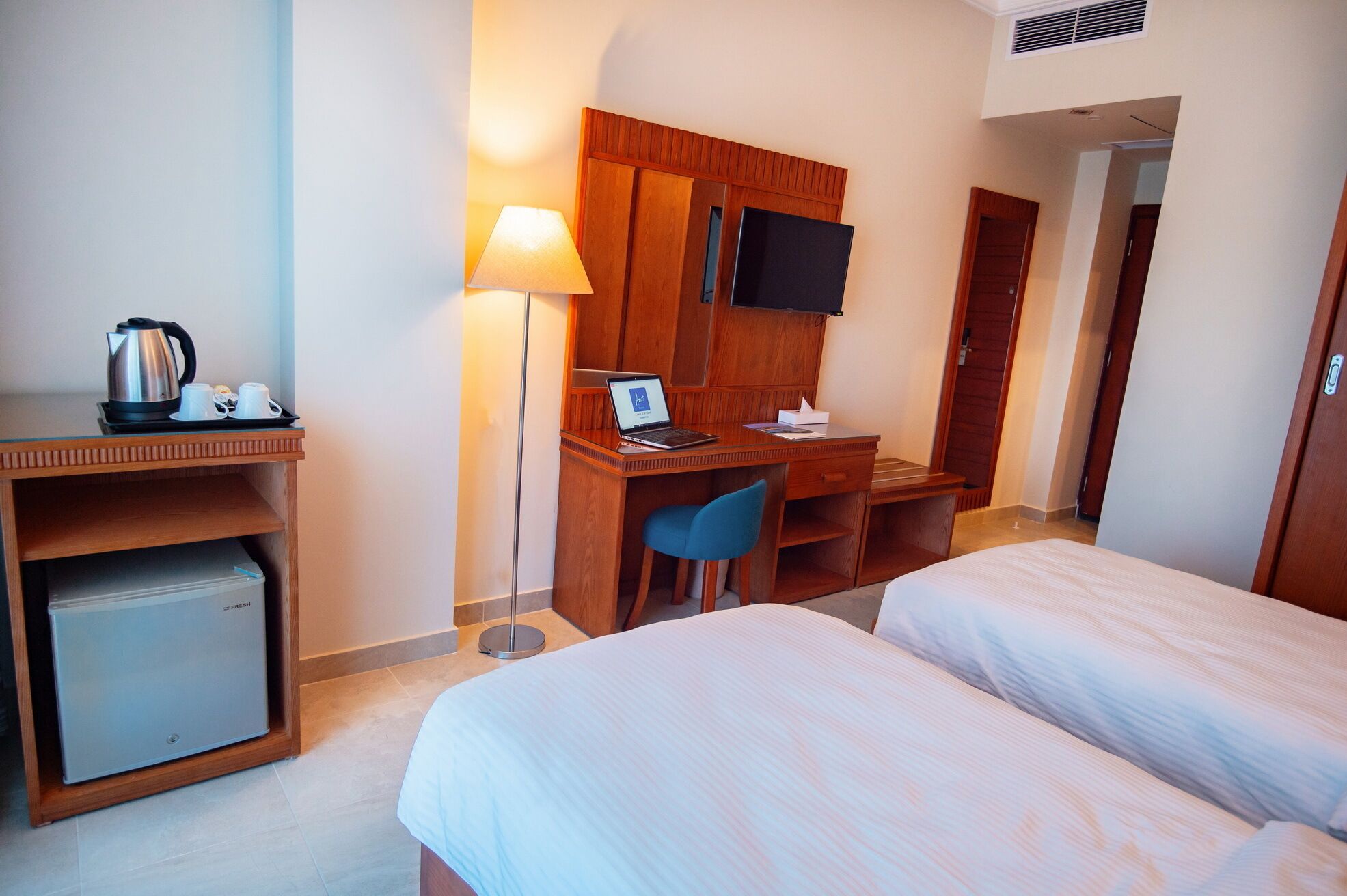 Habitación de hotel con dos camas, un escritorio con una computadora portátil, un refrigerador pequeño, una cafetera y un televisor en la pared.