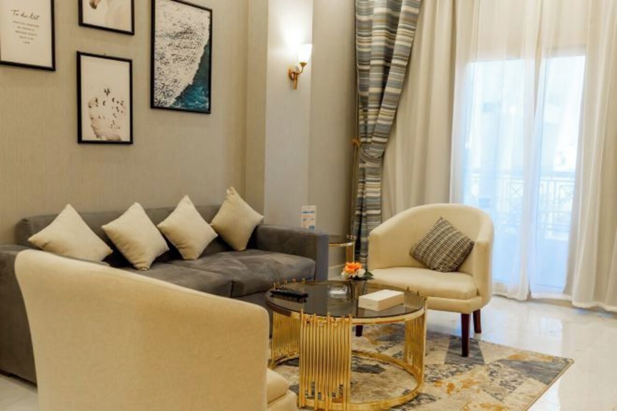 A modern living room with a grey sofa, a beige armchair, and a glass coffee table. There are framed pictures on the wall and a window with curtains.