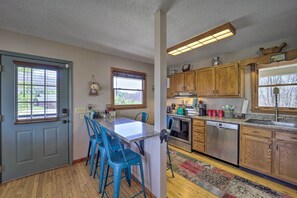 A kitchen with wooden cabinets, a granite countertop, and a built-in dishwasher. There are two windows with blinds, a door, and a rug on the floor. - Walk to New River Access! Family Cabin w/ Deck (Piney Creek)