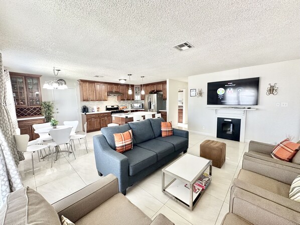 Una sala moderna con un sofá gris, una mesa de centro blanca y sillones marrones. Se ve una cocina con muebles de madera y un comedor con sillas blancas y una mesa. Un televisor de pantalla plana está montado en la pared.