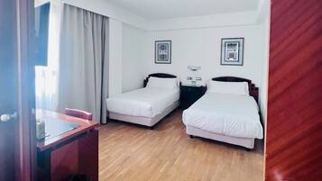 Habitación de hotel con dos camas individuales, un escritorio con silla, un pequeño cubo de basura y una lámpara en el techo.
