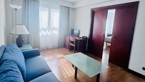 A modern living room with a blue sofa, a glass coffee table, and a wooden floor. There is a desk with a chair and a television in the background. - Hotel ITC Ciudad de Haro (Haro)