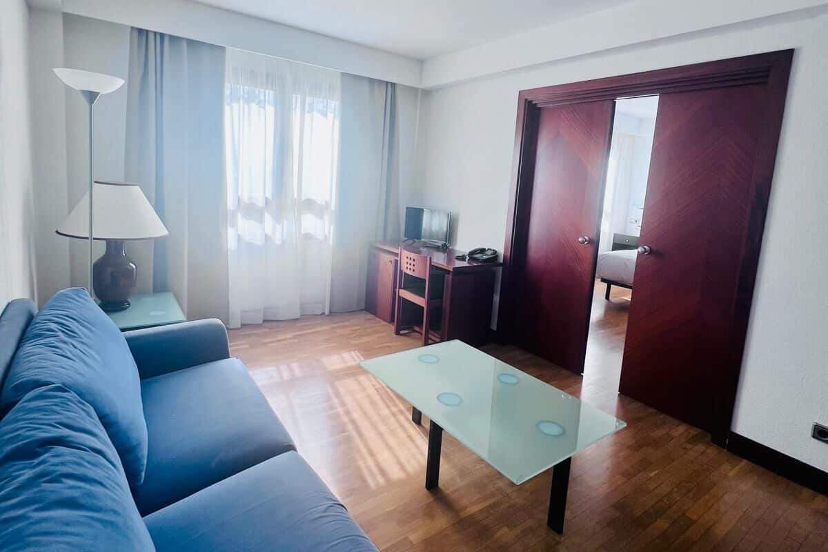 Un salon moderne avec un canapé bleu, une table basse en verre et un plancher en bois. On aperçoit un bureau avec une chaise et un téléviseur au fond de la pièce.