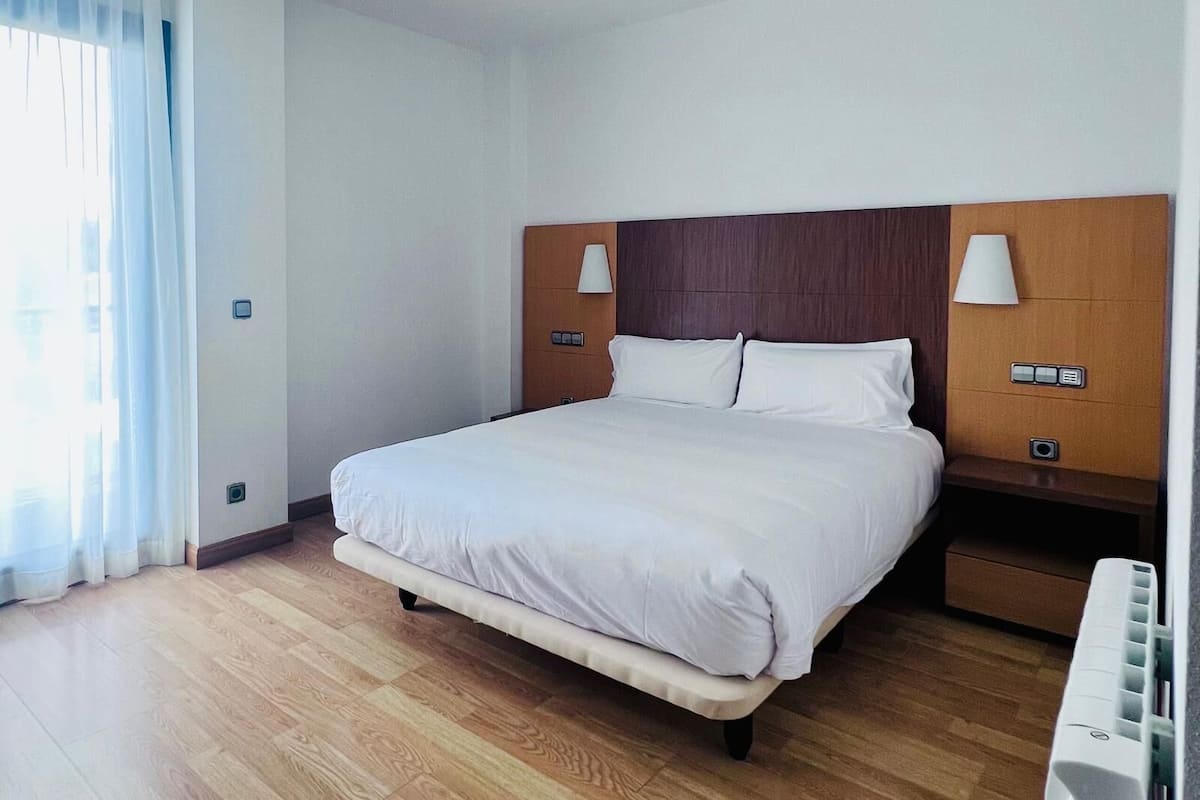 Une chambre d’hôtel moderne avec un grand lit, une tête de lit en bois, des lampes de chevet et une grande fenêtre avec des rideaux.