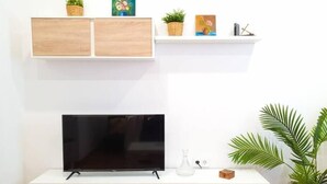 A modern living room with a flat-screen TV on a white console, wooden cabinets below, and a shelf above with decorative items and plants. - Casa Marialuisa - Casa original en el Casco antiguo (Barbate)