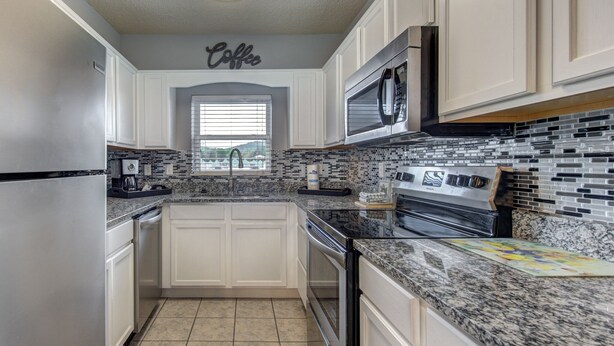 A kitchen with white cabinets, a granite countertop, a stainless steel refrigerator, a microwave, and a tiled backsplash.