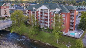 A multi-story hotel with a swimming pool, located near a river and surrounded by greenery and mountains.