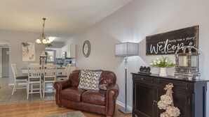 A living room with a brown leather sofa, a dining area with white chairs, a kitchen in the background, and a wall with a 'Welcome' sign.
