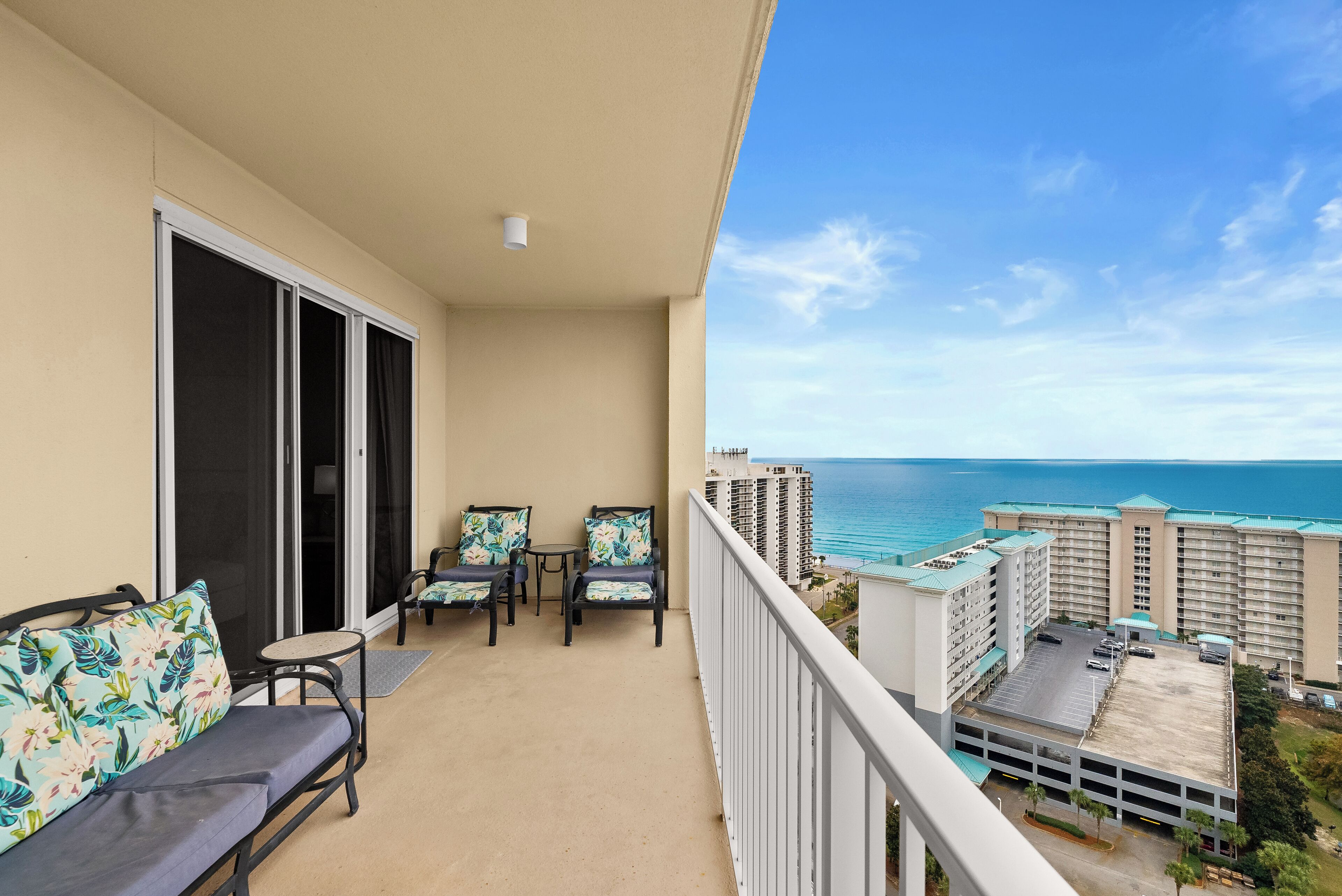 A balcony with a sofa, chairs, and a table, overlooking a parking area and a view of the ocean.
