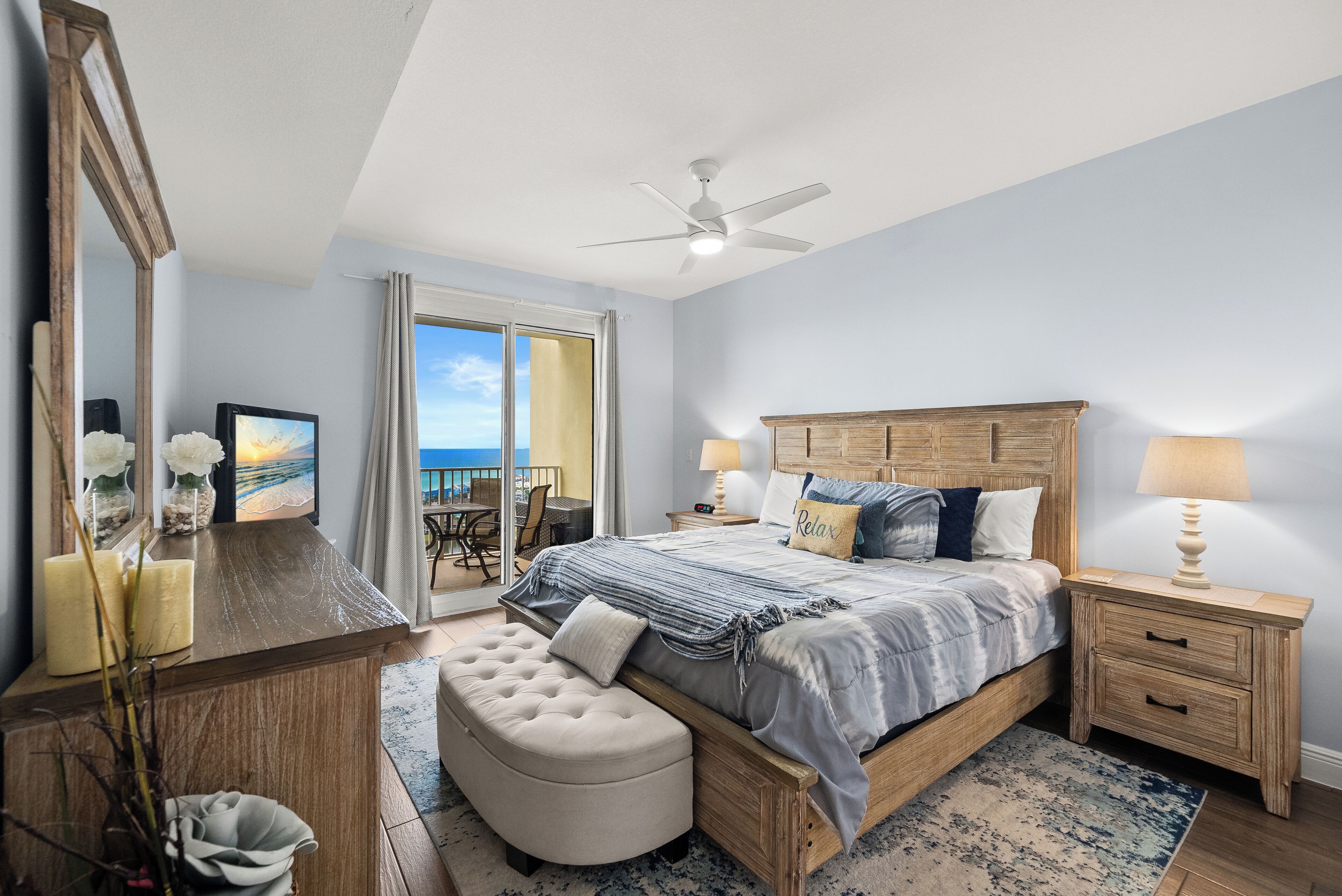 A bedroom with a large bed, a wooden dresser, a vanity with a mirror, and a view of the outdoors.