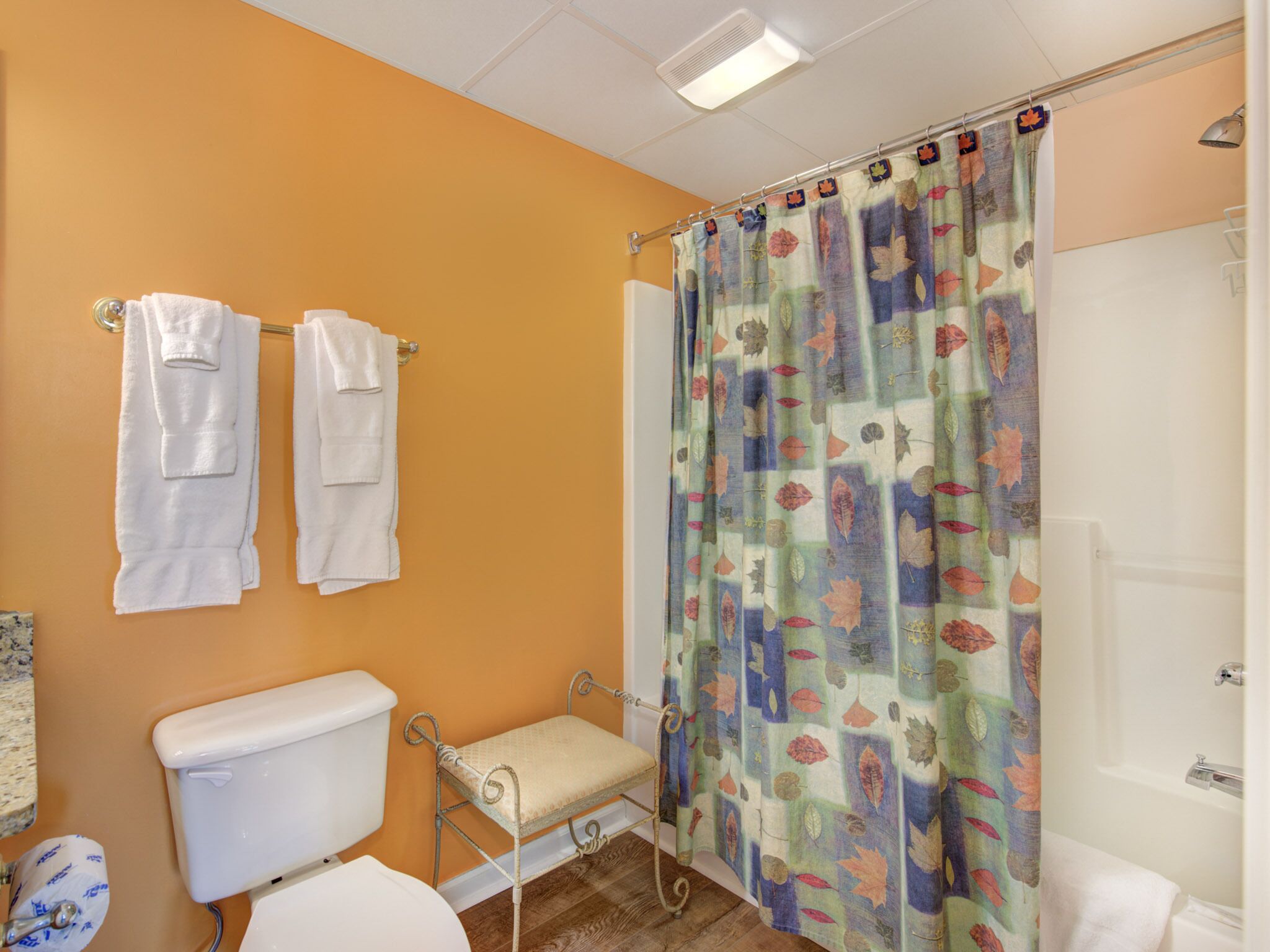 Un bagno con una parete arancione, una tenda da doccia con motivo a foglia e due asciugamani bianchi appesi a un portaserviti.