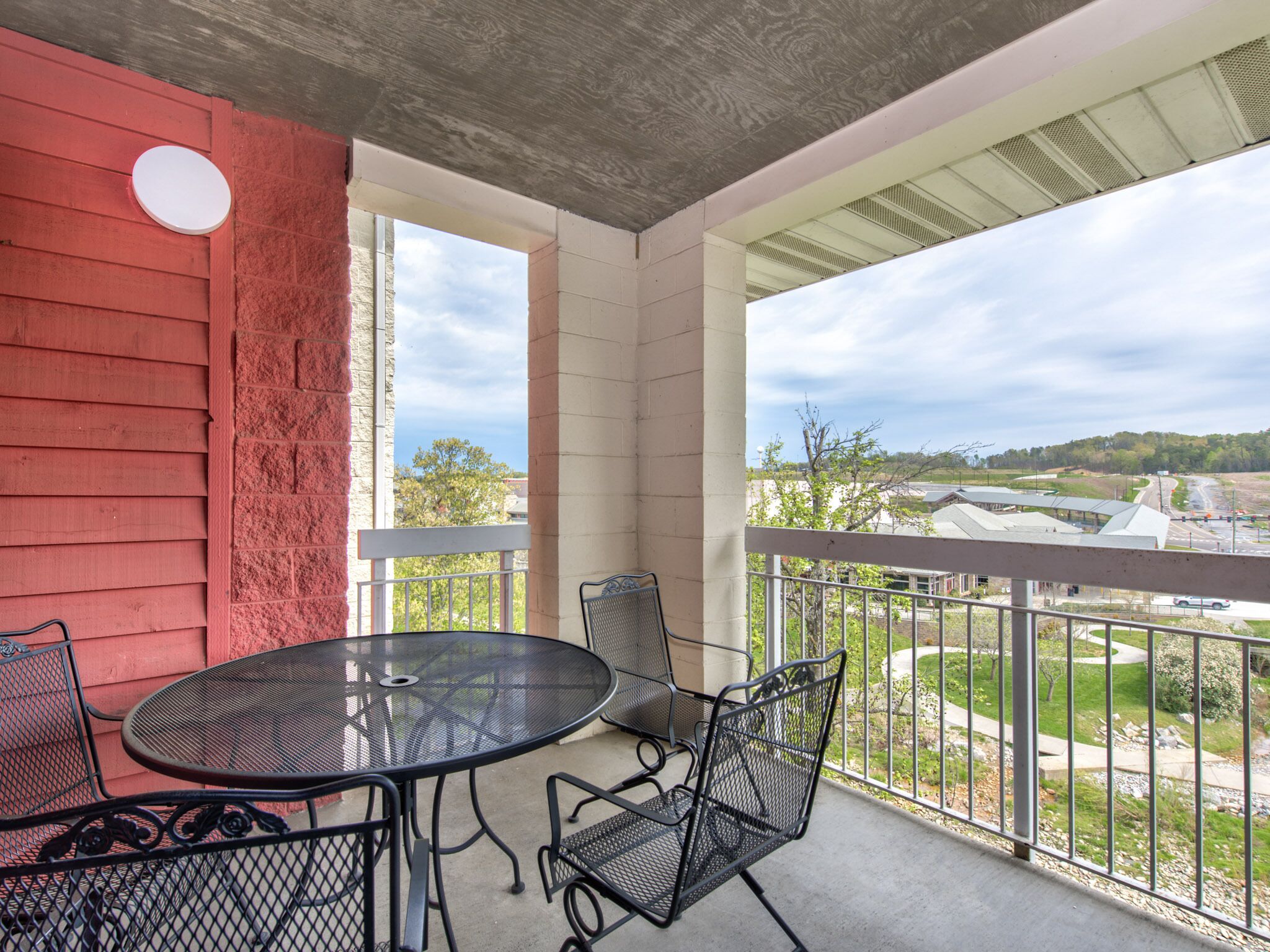 A balcony with a round table and chairs, a red wall, and a view of a road and greenery.