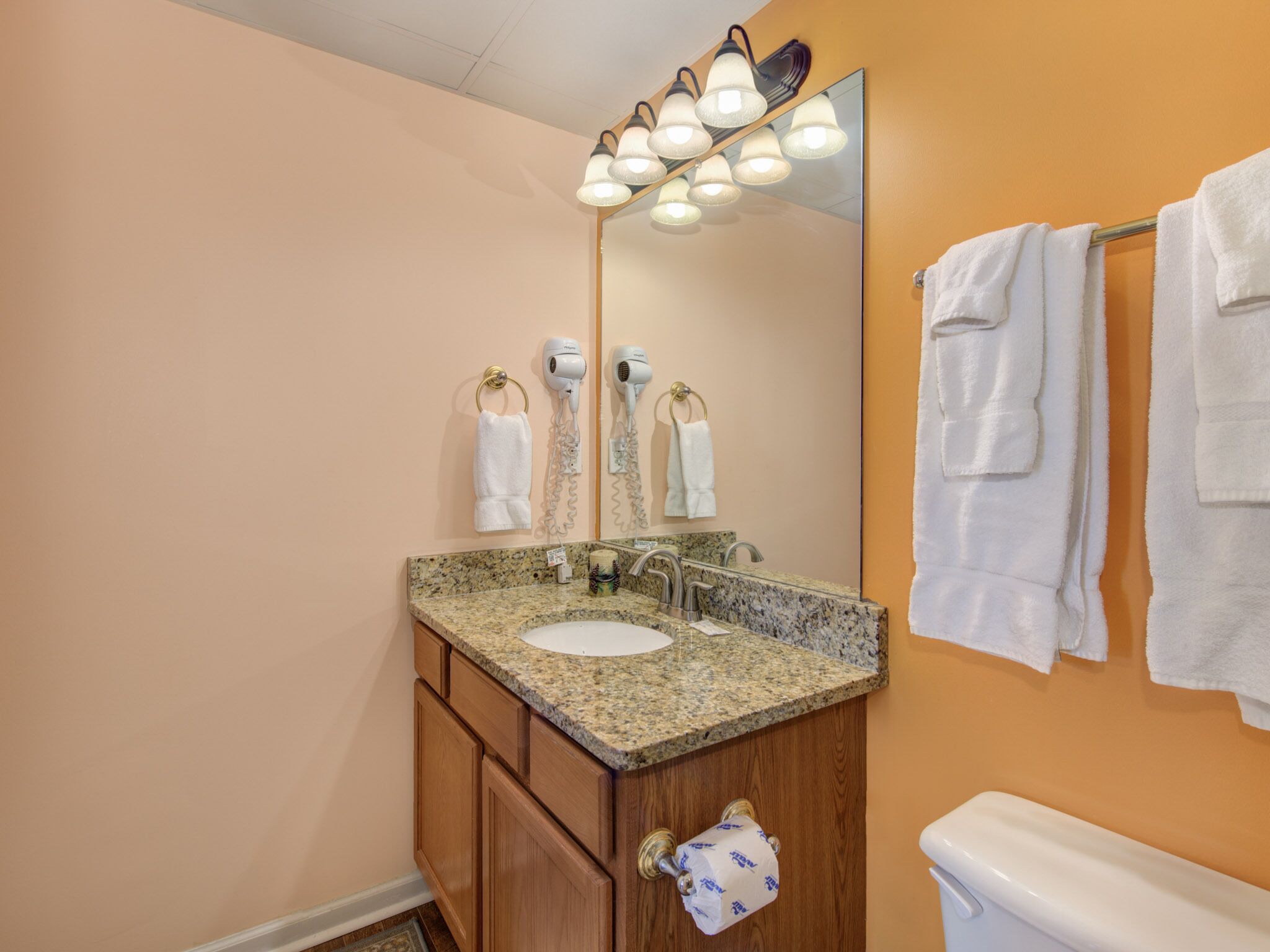 Une salle de bain avec un lavabo doté d’un plan de travail en granit, un miroir et des toilettes. Des serviettes sont suspendues sur un porte-serviettes et un sèche-cheveux est fixé au mur.