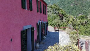 A pink building with green shutters, a stone pathway, and a mountainous backdrop.