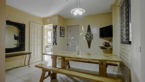 Un comedor con una mesa de madera y bancos, un lámpara del techo, un televisor montado en la pared y un adorno decorativo en la pared.