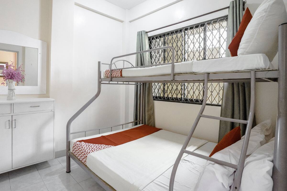 Una cama litera con estructura metálica, ropa de cama blanca y una manta marrón. Hay una mesita con un jarrón de flores y un espejo en la pared.