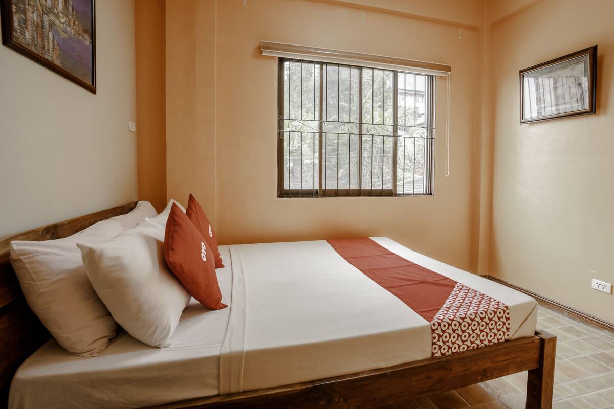 Una cama individual con estructura de madera, ropa de cama blanca y un corredor estampado. Se ve una ventana con barrotes y vistas a la vegetación.
