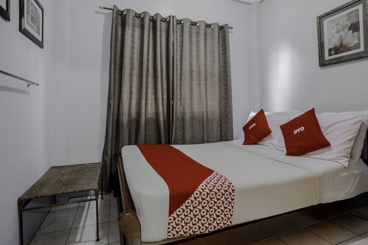Una cama bien hecha con una colcha estampada y dos almohadas, una mesita y una cortina.