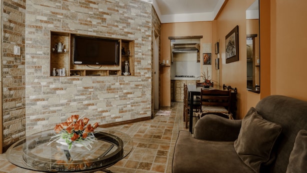 Una sala de estar con pared de ladrillo, una mesa de centro de vidrio con flores, un sofá marrón y una cocina al fondo.