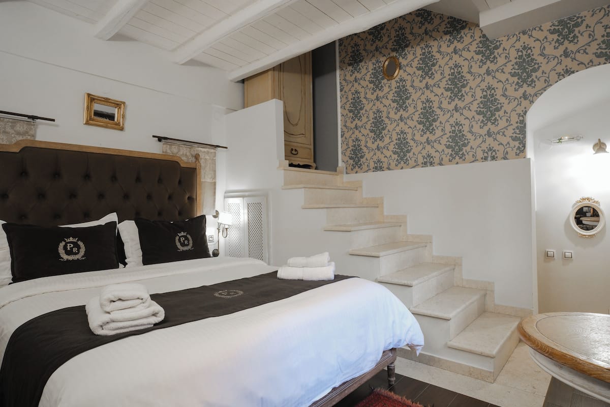 A bedroom with a large bed, a headboard, a staircase, and a bathroom with a round mirror.