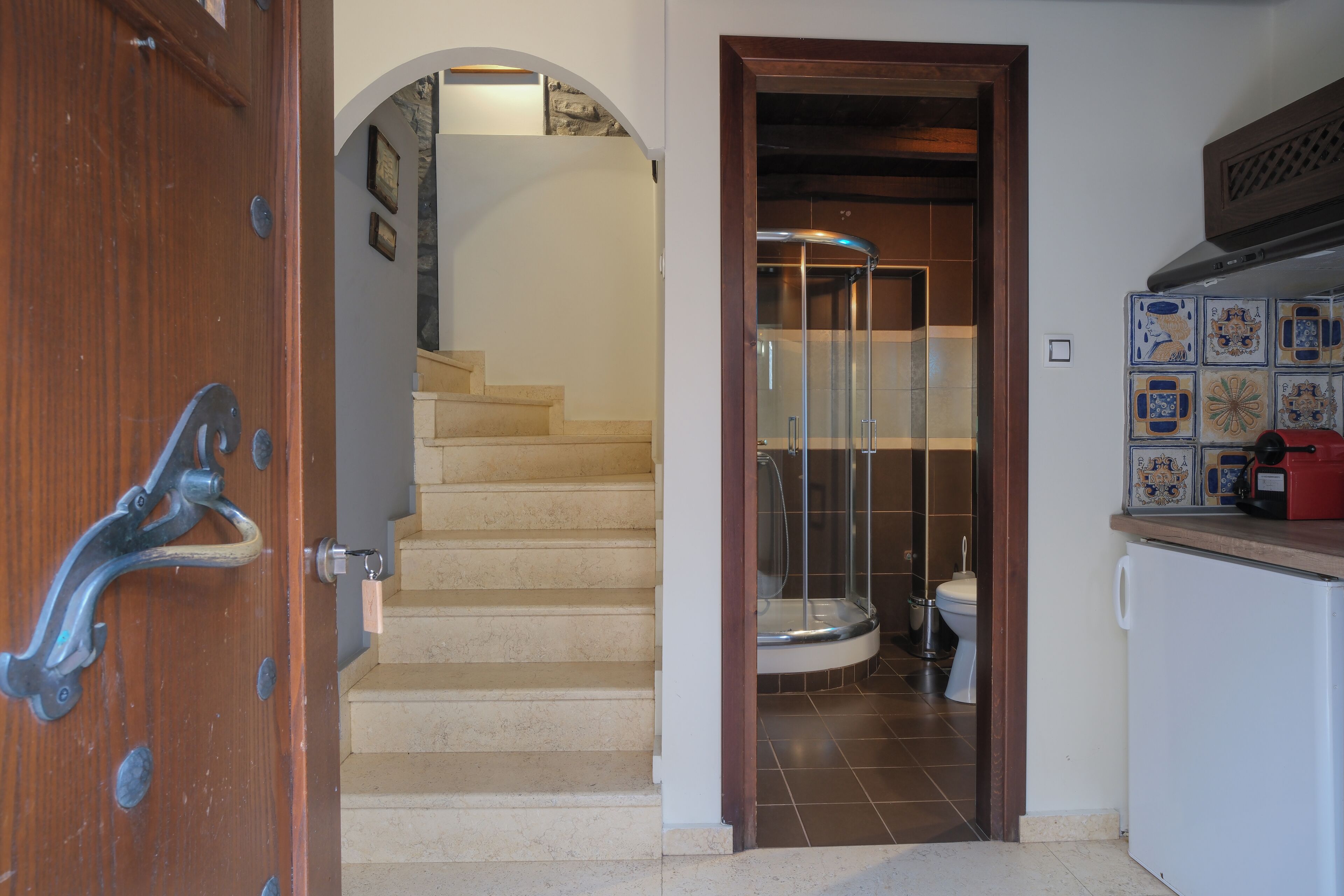 Una porta in legno con maniglia metallica, una scala che conduce al bagno con box doccia in vetro e un angolo cottura con frigorifero e piastrelle decorative.