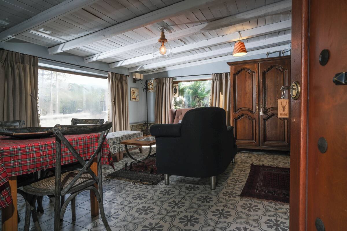 A rustic room with a wooden ceiling, a dining table with a red plaid tablecloth, a black armchair, and a wooden cabinet.