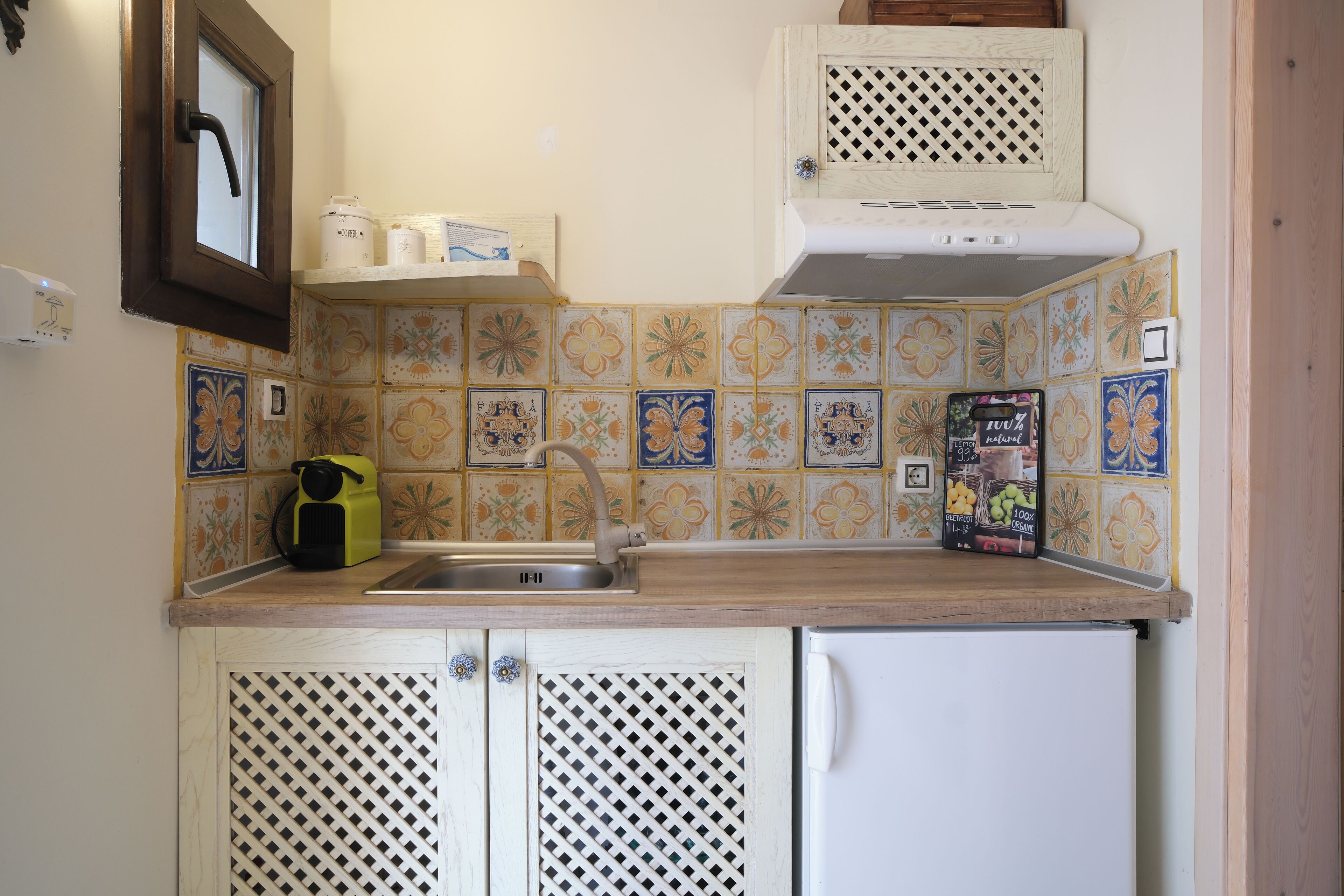 A kitchen with a sink, a white refrigerator, and a microwave on the countertop. There are decorative tiles on the backsplash and a window with a towel hanging on the door.