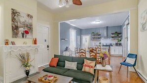 A spacious living room with a green sofa, a coffee table, and a fireplace. There is a kitchen in the background with white cabinets and a dining area.