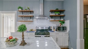 A kitchen with white cabinets, a marble countertop, a gas stove, a microwave, and shelves with plants and kitchenware.