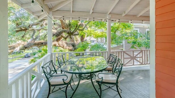 A covered outdoor area with a round glass table and chairs, surrounded by a white railing, overlooking a lush garden with large trees.