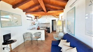 A living room with a blue sofa, a small table, and two chairs. There is a kitchen in the background with white cabinets and a wooden ceiling. - Terza Spiaggia la Filasca (Golfo Aranci)