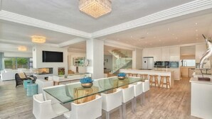 A spacious living area with a glass dining table, white chairs, and a modern kitchen in the background. - Marlin Manor (Struisbaai)