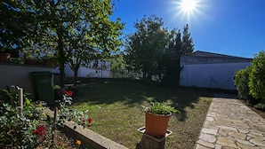 Een tuin met een stenen pad, een houten paal met een plant in een pot, en een groene prullenbak.