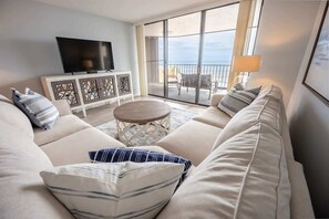 Een moderne woonkamer met een beige bank, een flatscreen televisie, een klein rond tafeltje en een balkon met uitzicht op zee.