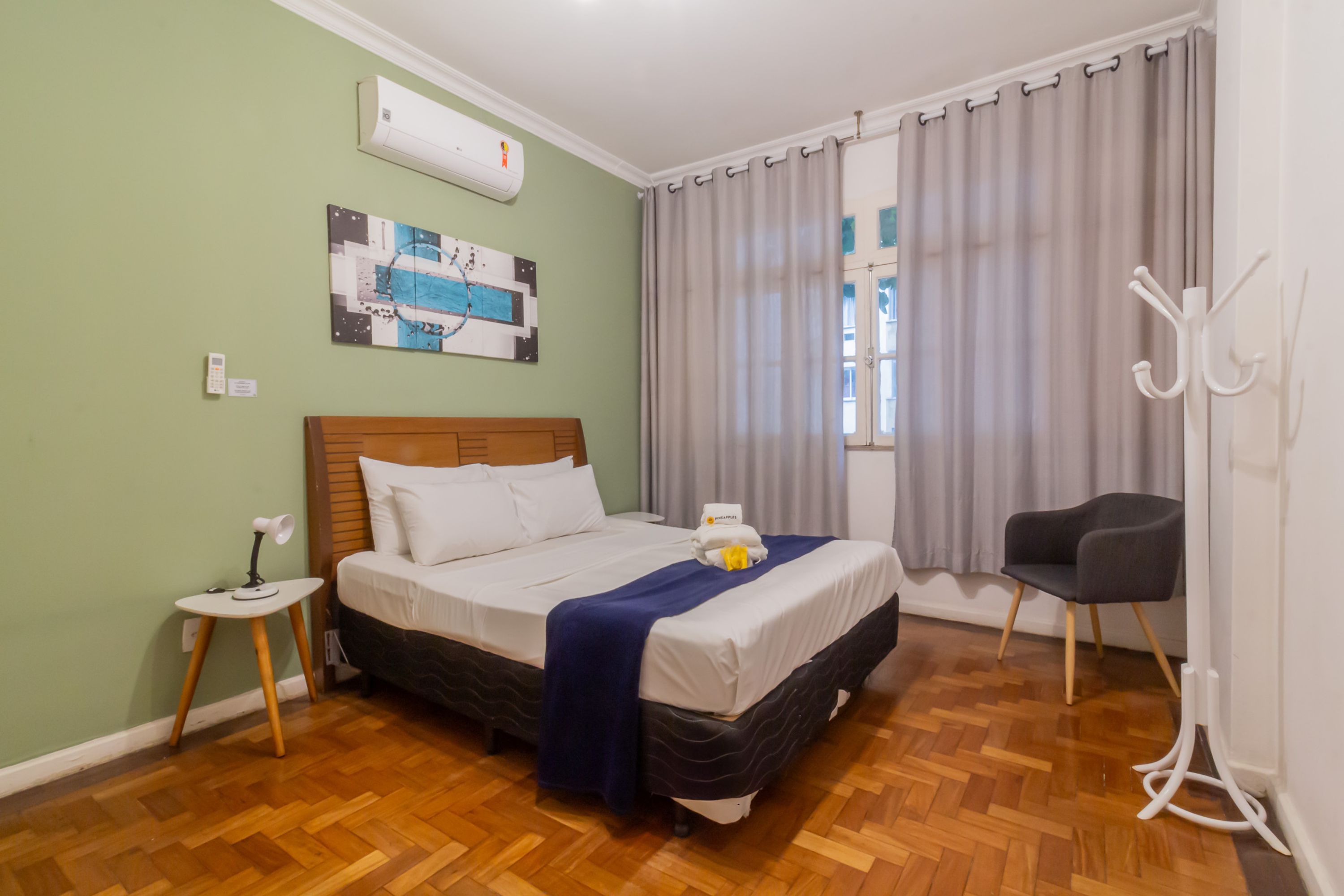A neatly arranged hotel room with a wooden floor, a bed with white bedding, a small bedside table, a chair, and a window with curtains.