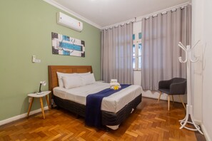 Een keurig opgemaakte hotelkamer met houten vloer, een bed met witte beddengoed, een klein nachtkastje, een stoel en een raam met gordijnen.