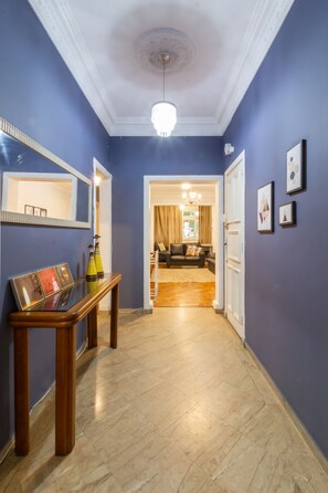 Een gang met een houten console tafel, ingelijste foto's en een spiegel. Deze leidt naar een zithoek met een bank en een kroonluchter.