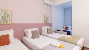 Een hotelkamer met drie eenpersoonsbedden, elk met witte beddengoed en diverse kussens. Er is een klein tafeltje met een stoel, een raam met een blauw gordijn en een airconditioner aan de muur.