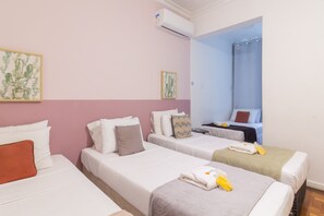 Een hotelkamer met drie eenpersoonsbedden, elk met witte beddengoed en diverse kussens. Er is een klein tafeltje met een stoel, een raam met een blauw gordijn en een airconditioner aan de muur.