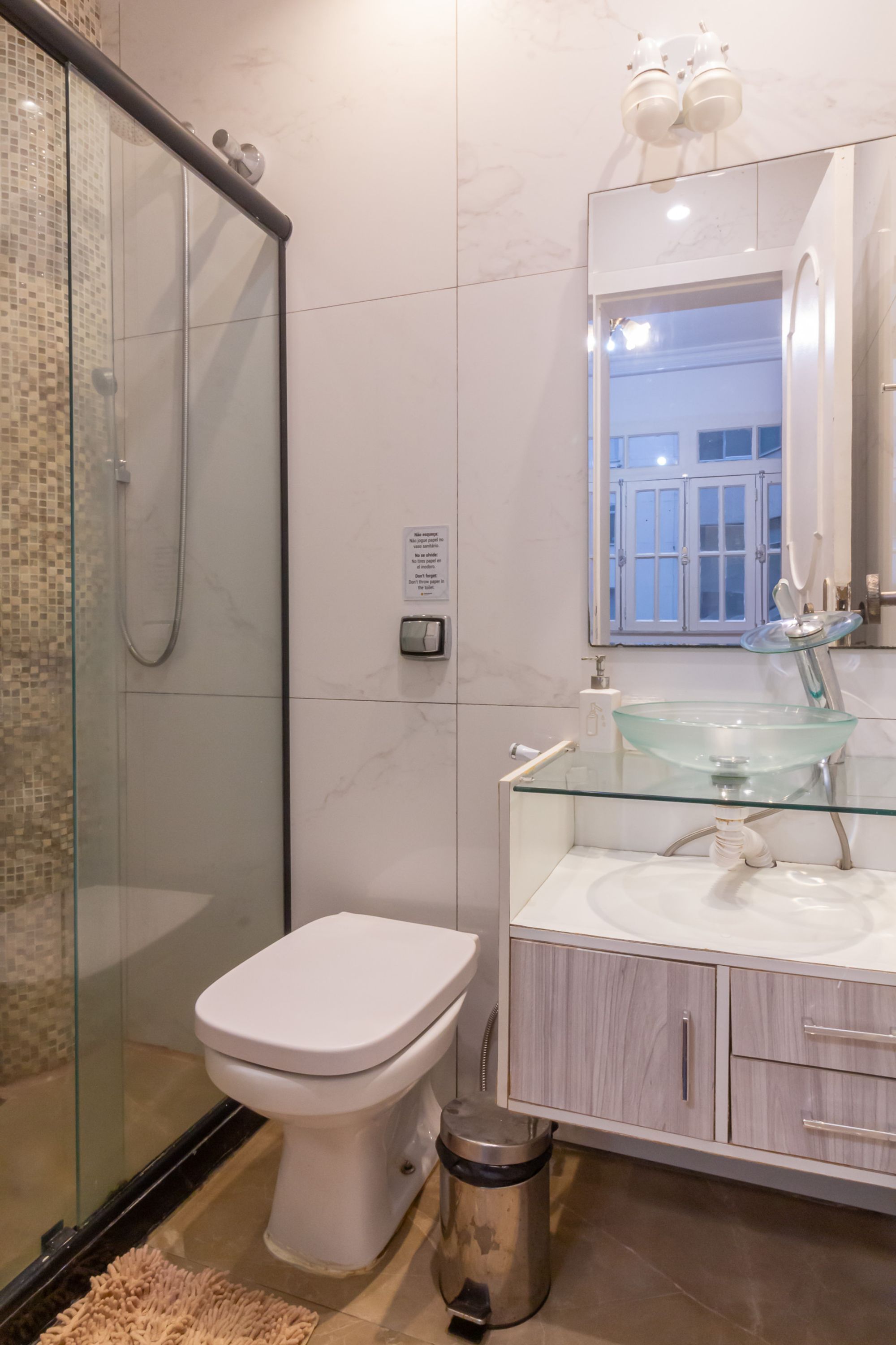 Un baño moderno con cabina de ducha de vidrio, inodoro blanco, lavamanos de vidrio y encimera de mármol, y tocador de madera.